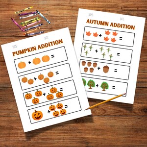 May include: Two white worksheets with fall-themed addition problems, including pumpkins, leaves, and acorns. The worksheets are titled "Pumpkin Addition" and "Autumn Addition." Crayons and a pencil are on a wooden surface.