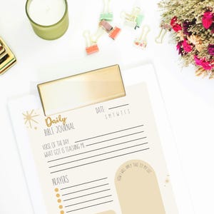 May include: A Daily Bible Journal with a gold clip, featuring prompts for reflection. The journal is accompanied by a gold stapler, colourful binder clips, a candle, and a bouquet of dried flowers. The journal's text includes "Verse of the Day" and "Prayers."