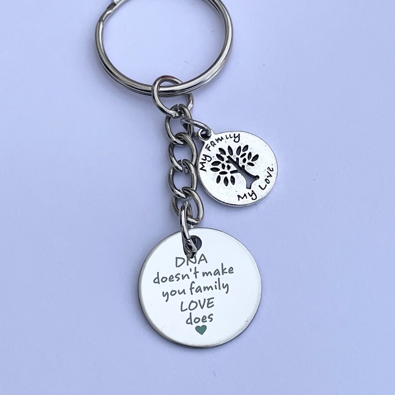 May include: Silver keychain with two round charms. The larger charm has the text "DNA doesn't make you family LOVE does" with a small green heart. The smaller charm has a tree silhouette and the text "My Family My Love".