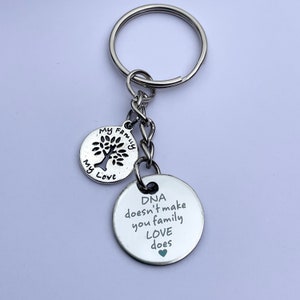 May include: Silver keychain with two round charms. The first charm has a tree cutout and the words "My Family My Love". The second charm has the text "DNA doesn't make you family LOVE does" with a small green heart.