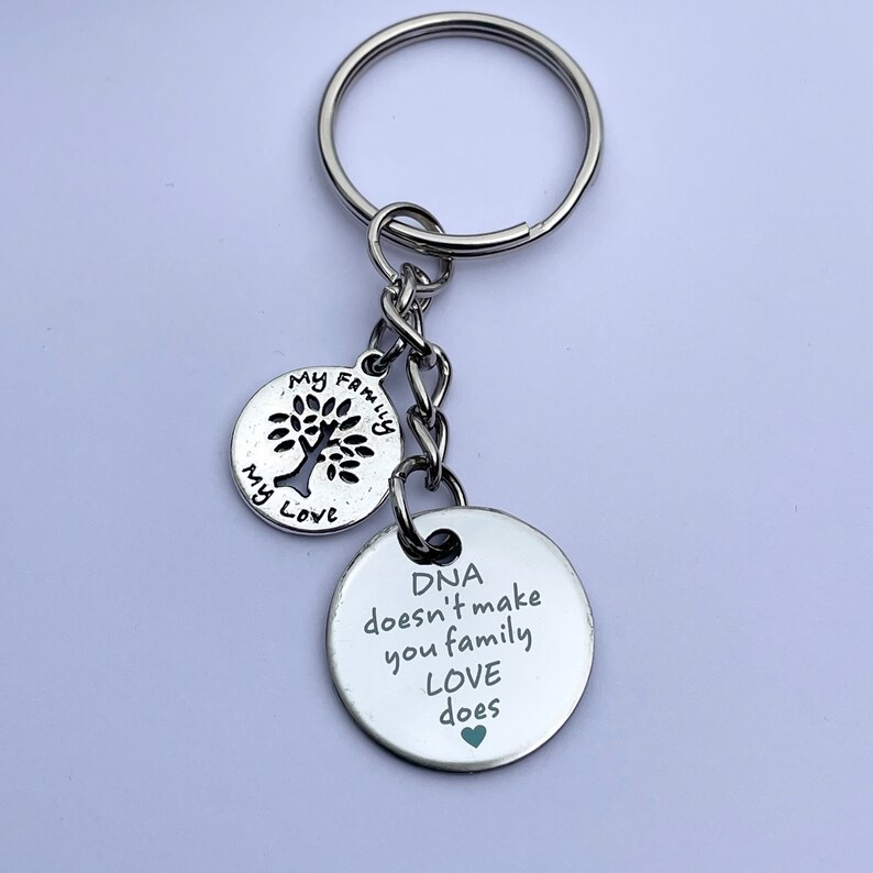 May include: Silver keychain with two round charms. The first charm has a tree cutout and the words "My Family My Love". The second charm has the text "DNA doesn't make you family LOVE does" with a small green heart.