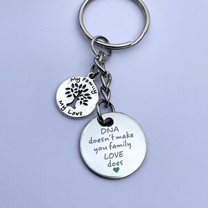 May include: Silver keychain with two round charms. The larger charm has the text "DNA doesn't make you family LOVE does" with a small green heart. The smaller charm has a tree with the text "My Family My Love".
