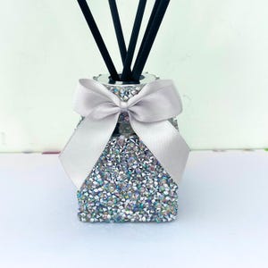 May include: A silver diffuser bottle with a white bow, covered in iridescent rhinestones. Black diffuser sticks are in the bottle.