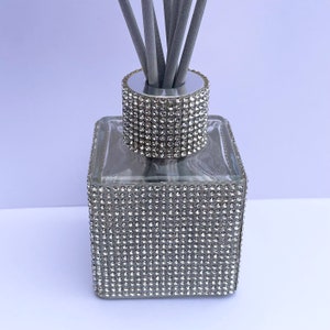 May include: A square glass diffuser bottle with a silver lid and a silver base. The bottle is covered in small, clear rhinestones. The bottle has grey diffuser sticks.