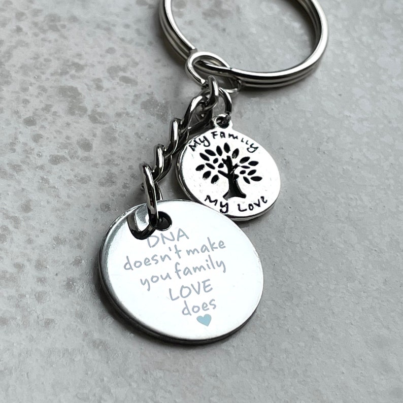 May include: Silver keychain with two round charms. The larger charm has the text "DNA doesn't make you family LOVE does" with a small blue heart. The smaller charm has a tree with the text "My Family My Love".
