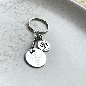 May include: Silver keychain with two charms. The larger charm has the text "DNA doesn't make you family LOVE does" with a small blue heart. The smaller charm has a tree silhouette and the text "My Family My Love".