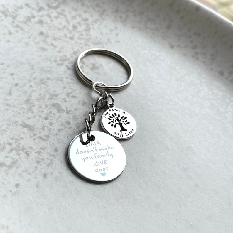 May include: Silver keychain with two charms. The larger charm has the text "DNA doesn't make you family LOVE does" with a small blue heart. The smaller charm has a tree silhouette and the text "My Family My Love".
