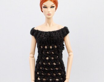 Puede incluir: Un vestido negro de ganchillo, con hombros descubiertos y aspecto texturizado. El vestido presenta patrones calados en el corpiño y la falda. El vestido se muestra en una muñeca con pelo rojo sobre un fondo blanco.