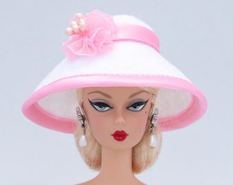May include: A white sun hat with a pink ribbon and a pink flower accent. The hat is worn by a doll with blonde hair and blue eyes.