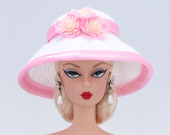 Könnte beinhalten: Ein weißer Sonnenhut mit einem rosa Band und zwei rosa Stoffblumen. Der Hut wird von einer Puppe mit blonden Haaren und Perlenohrringen getragen.