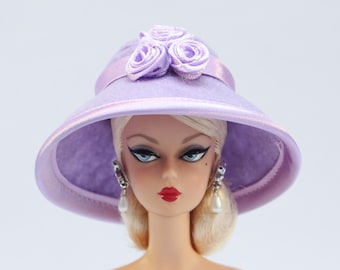 Könnte beinhalten: Eine Puppe mit einem lavendelfarbenen Hut mit breiter Krempe und drei dekorativen Stoffrosen. Die Puppe hat blondes Haar, blaue Augen und roten Lippenstift. Sie trägt auch Ohrringe mit Perltropfen.