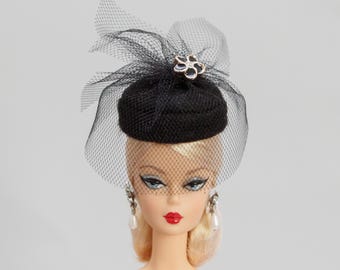 May include: A black doll-sized hat with a decorative flower and netting. The hat is perched on a doll's head, which is wearing pearl earrings and red lipstick. The doll has blonde hair and blue eyes.