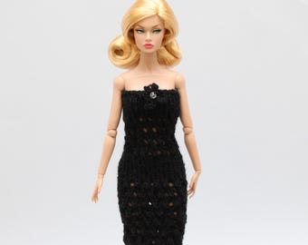 Puede incluir: Una muñeca de moda con un vestido negro tejido sin tirantes. El vestido tiene un pequeño elemento decorativo en el busto y una silueta ajustada. La muñeca tiene el pelo rubio peinado con una onda retro.