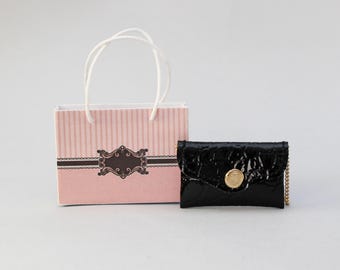 May include: A miniature black patent leather wallet with a gold-colored button and chain, next to a pink and white striped shopping bag with a decorative label.