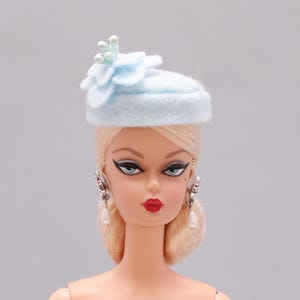 Puede incluir: Un sombrero de fieltro azul claro con un adorno de flor y detalles de perlas. El sombrero lo lleva una muñeca con el pelo rubio y los labios rojos.