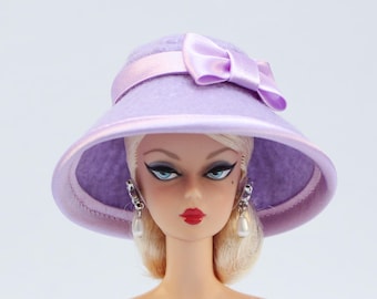 Könnte beinhalten: Eine Puppe mit blonden Haaren, blauen Augen und rotem Lippenstift trägt einen lavendelfarbenen Hut mit passender Schleife und rosa Band. Die Puppe hat Perlenohrringe. Der Hut hat eine breite Krempe.