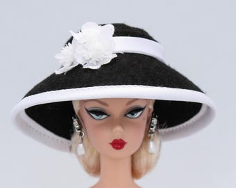 May include: A black felt hat with a white trim and a white flower accent. The hat is worn by a doll with blonde hair and blue eyes.