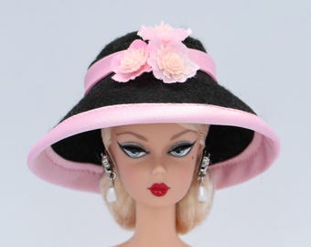 Könnte beinhalten: Ein schwarzer Filzhut mit einem rosa Band und rosa Blumenverzierungen. Der Hut hat eine breite Krempe und eine leicht abgerundete Krone.