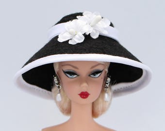 May include: A doll wearing a black hat with a white brim and a white ribbon. The hat is adorned with white flower accents. The doll has blonde hair, red lipstick, and pearl earrings. The doll is a fashion item.