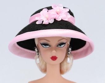 Könnte beinhalten: Ein schwarzer Filzhut mit rosa Satinbesatz und rosa Stoffblumen. Der Hut wird von einer Puppe mit blonden Haaren und blauen Augen getragen.