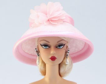 Könnte beinhalten: Eine Puppe mit blonden Haaren, blauen Augen und rotem Lippenstift, die einen hellrosa Hut mit einer großen Blume und Perlenohrringen trägt. Der Hut hat eine breite Krempe und ein Satinband.