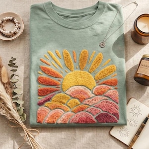May include: A sage green t-shirt with an embroidered sunrise design in yellow, orange, and pink. The shirt is displayed with a necklace, a candle, a bracelet, a small notebook, and dried flowers.