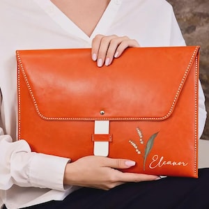 May include: Orange leather portfolio with a white strap and decorative stitching. The portfolio features a floral design and the name "Eleanor" in script. The portfolio is held by a person wearing a white shirt.