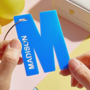 May include: A bright blue acrylic letter M tag featuring the name MADISON engraved vertically in white capital letters. The tag has a small hole at the top left corner with a thin metallic wire loop attached for hanging purposes.