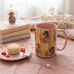 May include: A pink-handled coffee mug with a repeating pattern of hugging monkeys. A small monkey figurine is displayed under a glass cloche. A pink plate holds a white macaron and dried strawberries. Books and a spoon are also present.