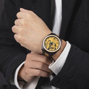 Op de afbeelding: Een zwart en houtkleurig polshorloge met een aangepaste foto op de wijzerplaat. Het horloge heeft een zwarte lunette en een band met afwisselende houten en zwarte schakels. De wijzerplaat toont een familieportret.