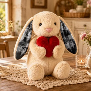 May include: A plush beige rabbit toy with long, floppy ears featuring black and white photo collages of people. The rabbit has a soft, textured surface, a small pink nose, and is holding a bright red, heart-shaped plush pillow in its front paws.