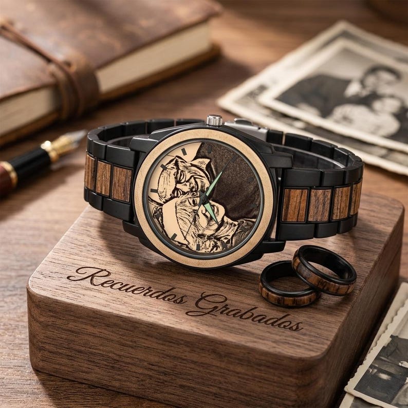 Op de afbeelding: Een houten horloge met een zwarte bezel en band, met een lichtgekleurde wijzerplaat met een gegraveerde afbeelding. Het horloge rust op een houten doos met de gegraveerde tekst "Recuerdos Grabados". Twee bijpassende ringen zijn ook zichtbaar.