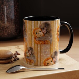 May include: A ceramic coffee mug with a black interior and handle. The exterior features a repeating pattern of a baby monkey hugging a stuffed orangutan. The background has vertical stripes in shades of yellow and beige.