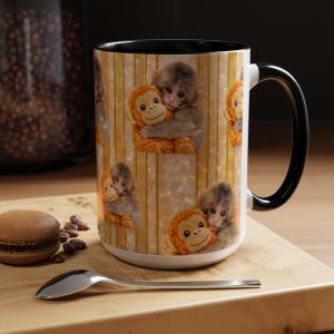 May include: White ceramic mug with a black handle and rim. The mug features a repeating pattern of baby monkeys hugging stuffed orangutans against a striped background. A spoon and a macaron are on a wooden surface.