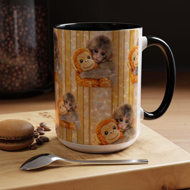 May include: White ceramic mug with a black handle and rim. The mug features a repeating pattern of baby monkeys hugging stuffed orangutans against a striped background. A spoon and a macaron are on a wooden surface.