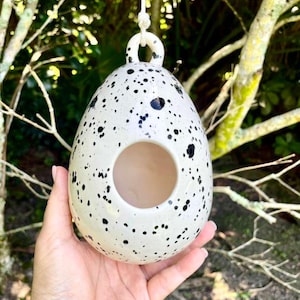 May include: An egg-shaped ceramic birdhouse with a glossy off-white finish and black speckled pattern. The structure features a circular opening on the front and a small loop at the top for hanging with an attached white cord for suspension.