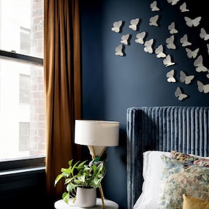 May include: A bedroom interior featuring a dark blue wall adorned with numerous white butterfly wall decals. A blue ribbed headboard sits below the butterflies, accompanied by a white lamp, a green potted plant, and brown curtains.