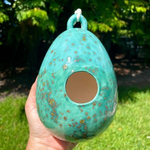 May include: An egg-shaped ceramic birdhouse with a glossy turquoise glaze featuring speckled brown and gold accents. The structure has a circular opening in the center and a small loop at the top with a white cord attached for hanging purposes.