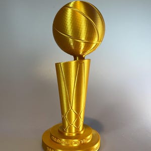 May include: A gold-coloured trophy featuring a basketball mounted on a tapered, net-patterned base. The circular pedestal is embossed with the text Oklahoma City Thunder 2025 NBA Champions, showcasing a metallic finish and a 3D-printed texture.