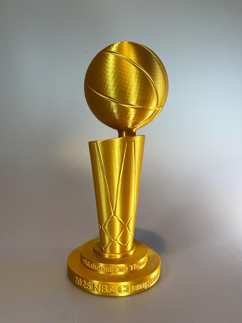 May include: A gold-coloured trophy featuring a basketball mounted on a tapered, net-patterned base. The circular pedestal is embossed with the text Oklahoma City Thunder 2025 NBA Champions, showcasing a metallic finish and a 3D-printed texture.