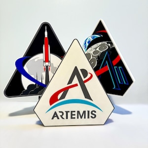 May include: Three triangular Artemis mission patches. One features a rocket launching, another shows the Earth and moon, and the third has the Artemis logo with the word "ARTEMIS" below. The patches have black, white, red, blue, and gray elements.
