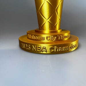 May include: A gold-coloured trophy base with a textured, diamond-patterned upper section. The base features embossed text reading Oklahoma City Thunder on the top tier and 2025 NBA Champions on the bottom tier, with a metallic, reflective finish.