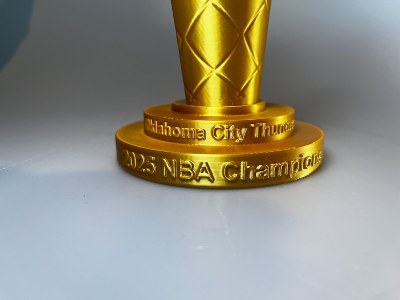 May include: A gold-coloured trophy base with a textured, diamond-patterned upper section. The base features embossed text reading Oklahoma City Thunder on the top tier and 2025 NBA Champions on the bottom tier, with a metallic, reflective finish.