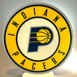 May include: A round sign featuring the Indiana Pacers logo. The logo includes a yellow circle with the team name, a navy blue "P" with a basketball, and a white background. The sign is mounted on a white stand.