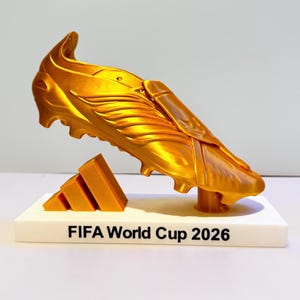 May include: A gold-colored 3D printed soccer cleat trophy mounted on a white rectangular base. The base features the black text "FIFA World Cup 2026" and a gold-colored three-stripe logo element positioned next to the cleat on the platform.