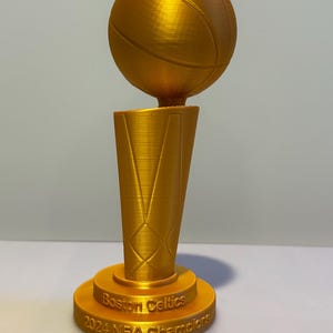 May include: A gold-coloured trophy featuring a basketball mounted on a tapered, textured base. The base is inscribed with the text Boston Celtics 2024 NBA Champions. The object has a visible 3D printed texture throughout its entire surface.