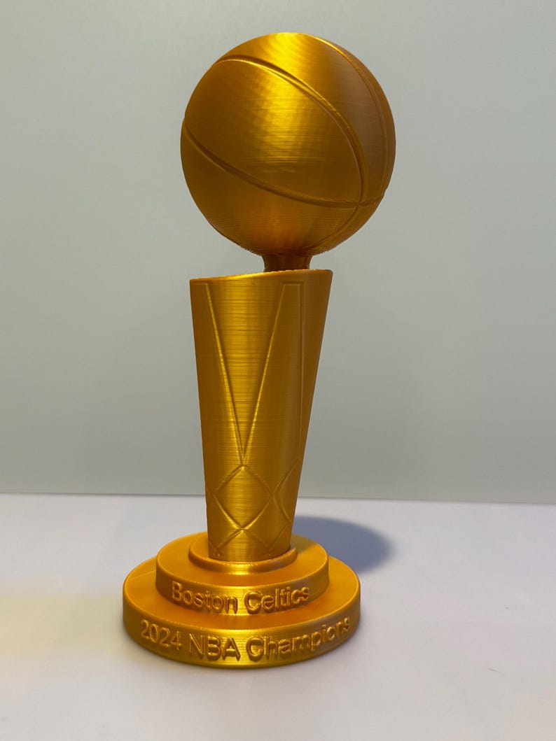 May include: A gold-coloured trophy featuring a basketball mounted on a tapered, textured base. The base is inscribed with the text Boston Celtics 2024 NBA Champions. The object has a visible 3D printed texture throughout its entire surface.
