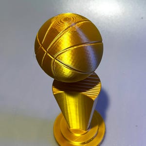 May include: A gold-coloured 3D printed trophy featuring a spherical basketball mounted on a tapered, cylindrical pedestal base. The object has a smooth, metallic finish with visible horizontal layer lines characteristic of additive manufacturing.