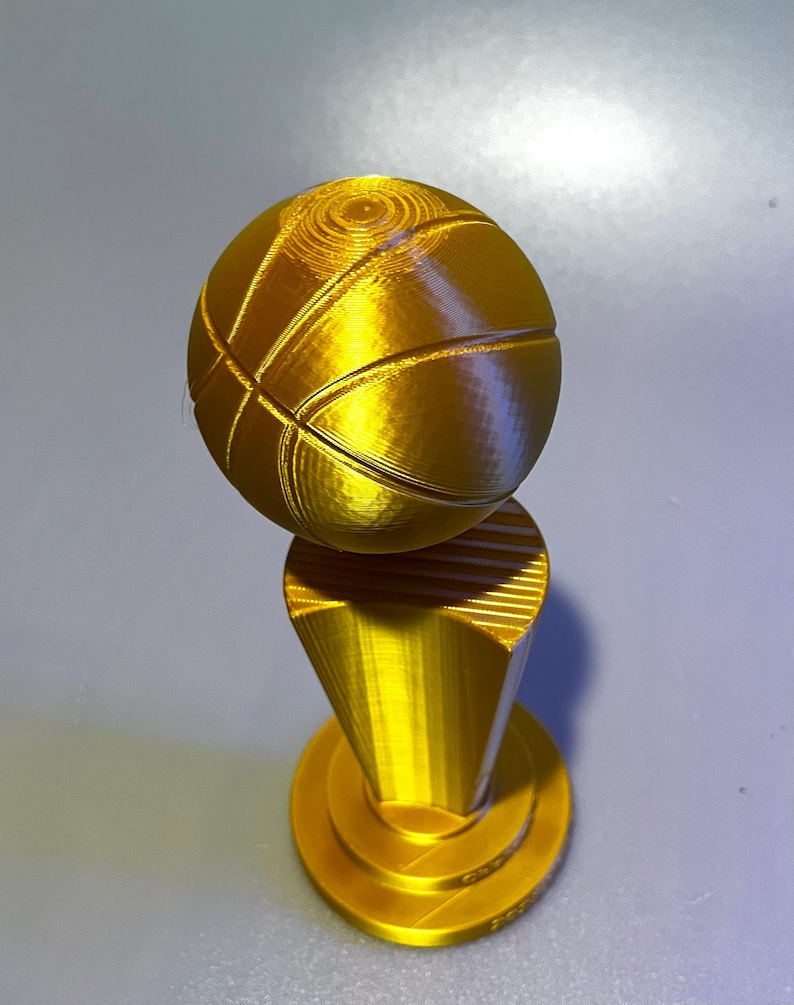 May include: A gold-coloured 3D printed trophy featuring a spherical basketball mounted on a tapered, cylindrical pedestal base. The object has a smooth, metallic finish with visible horizontal layer lines characteristic of additive manufacturing.