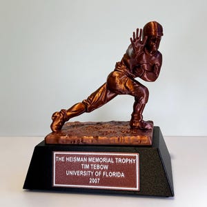 May include: A bronze-colored trophy depicting a football player in a stiff-arm pose on a textured base. The black pedestal features a plaque reading: THE HEISMAN MEMORIAL TROPHY, TIM TEBOW, UNIVERSITY OF FLORIDA, 2007. The figure is in a dynamic stance.